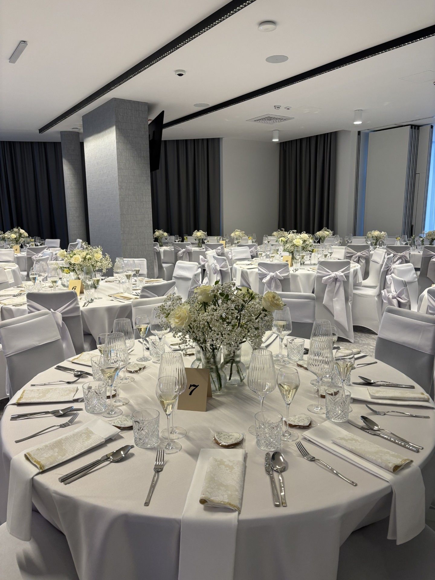 Uma sala de banquetes com mesas redondas preparadas para um evento formal, com toalhas brancas, copos e arranjos florais.