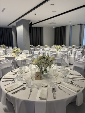 Un salón de banquetes con mesas redondas dispuestas para un evento formal, con manteles blancos, cristalería y centros de mesa florales.