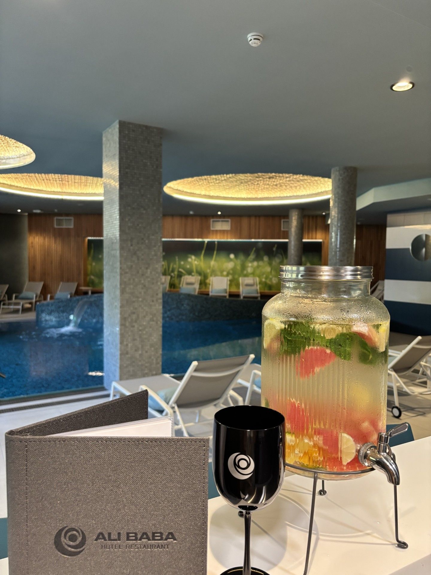 A hotel room with a view of a swimming pool, a large glass jar with a drink, and a black mug on a table.