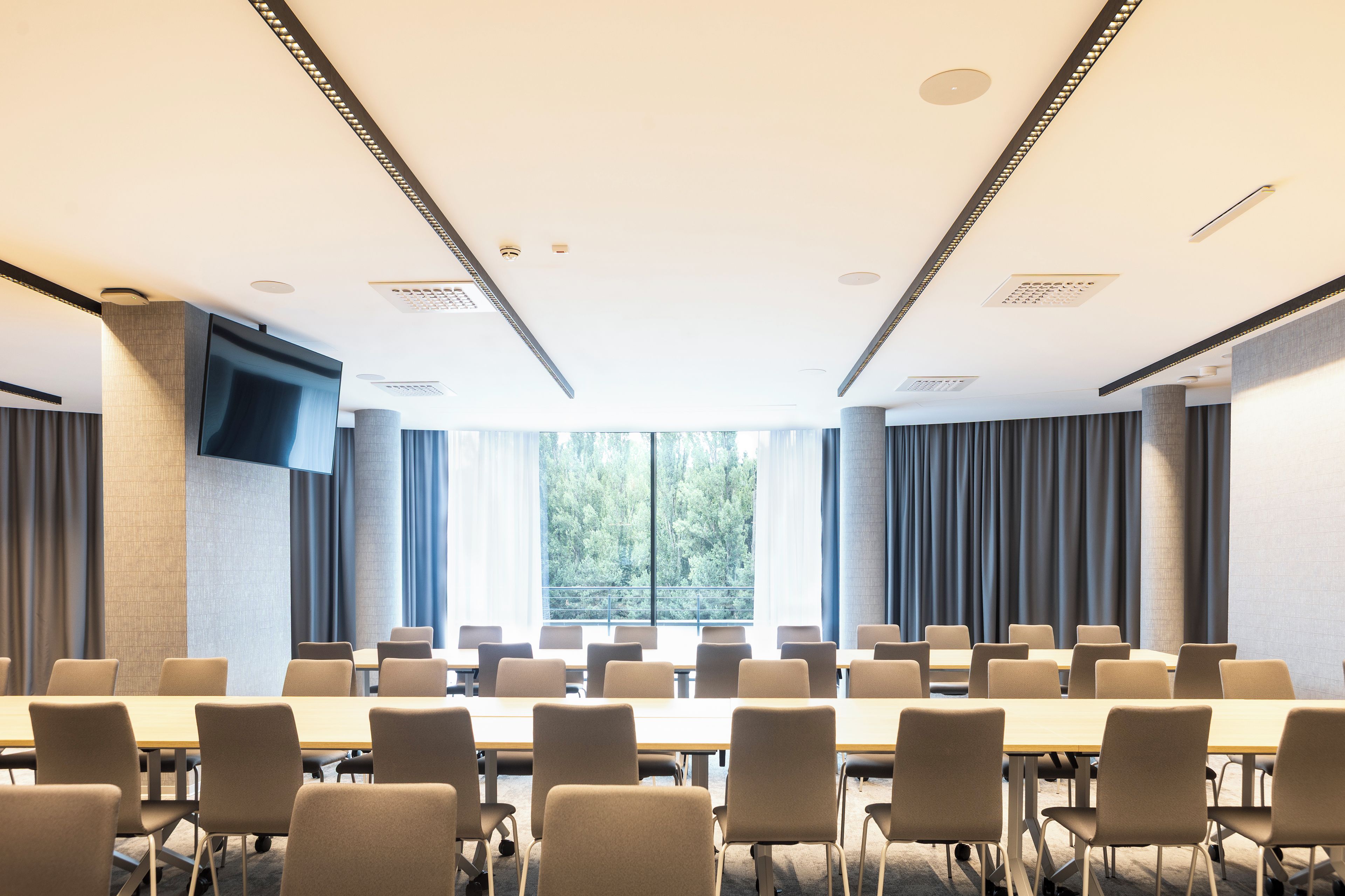 A conference room with a large table and chairs, a flat-screen TV mounted on the wall, and large windows with curtains.