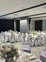 Un salón de banquetes con mesas redondas, sillas blancas y centros de mesa florales.