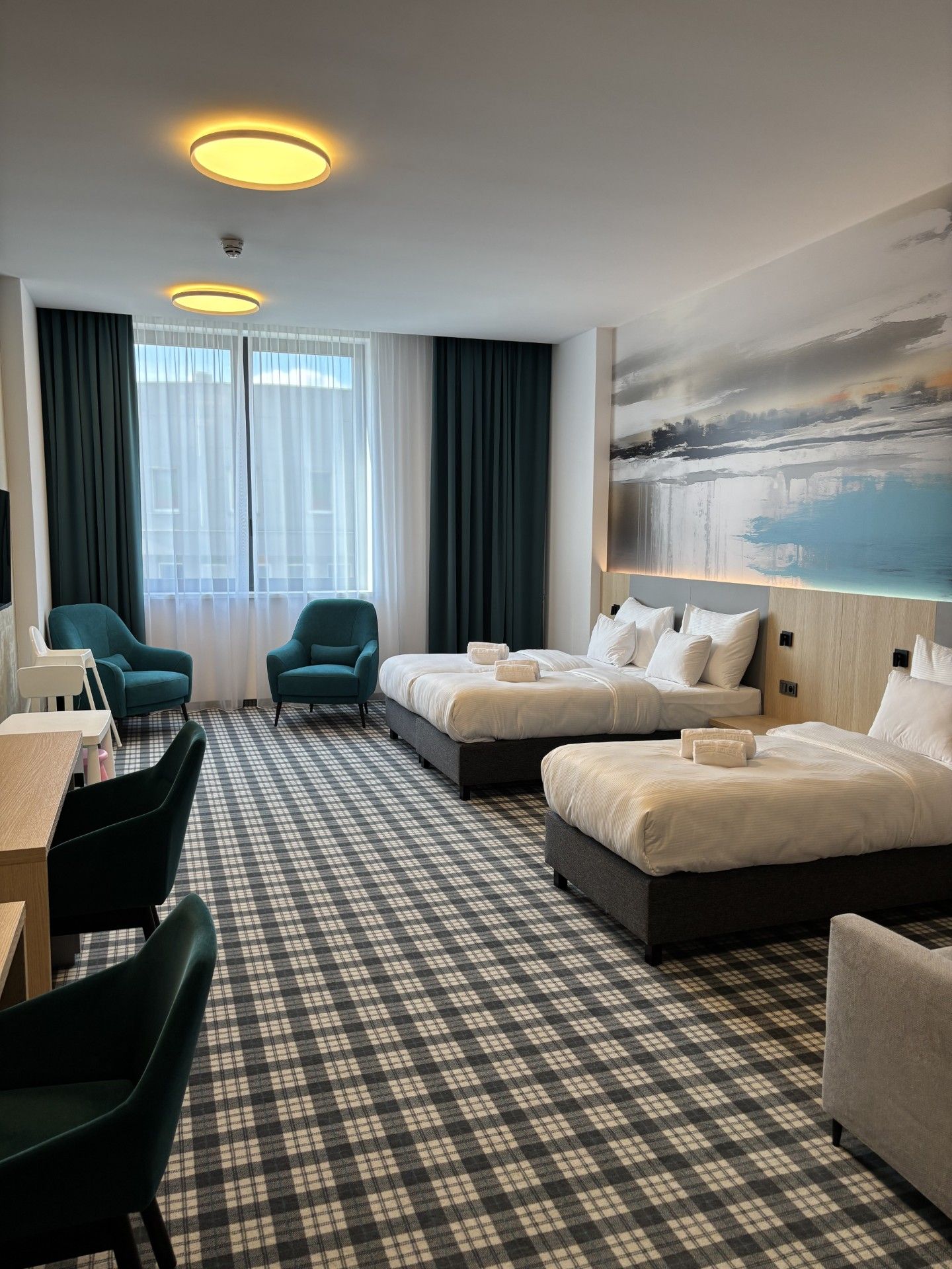 A hotel room with a checkered floor, two beds, a desk, chairs, and a painting on the wall.