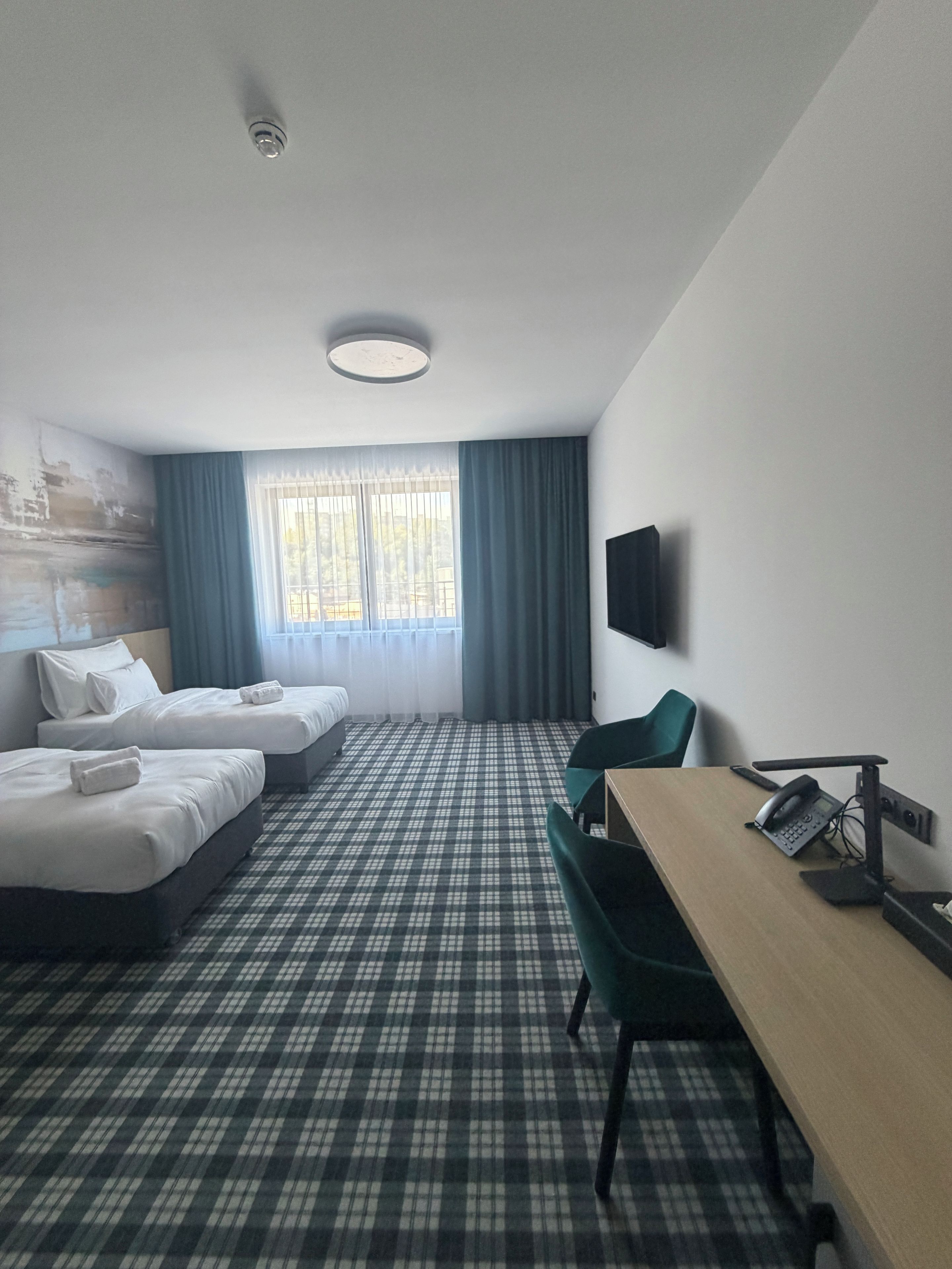 Ein Hotelzimmer mit zwei Betten, einem Schreibtisch, einem Stuhl, einem Flachbildfernseher und einem Fenster mit Vorhängen.