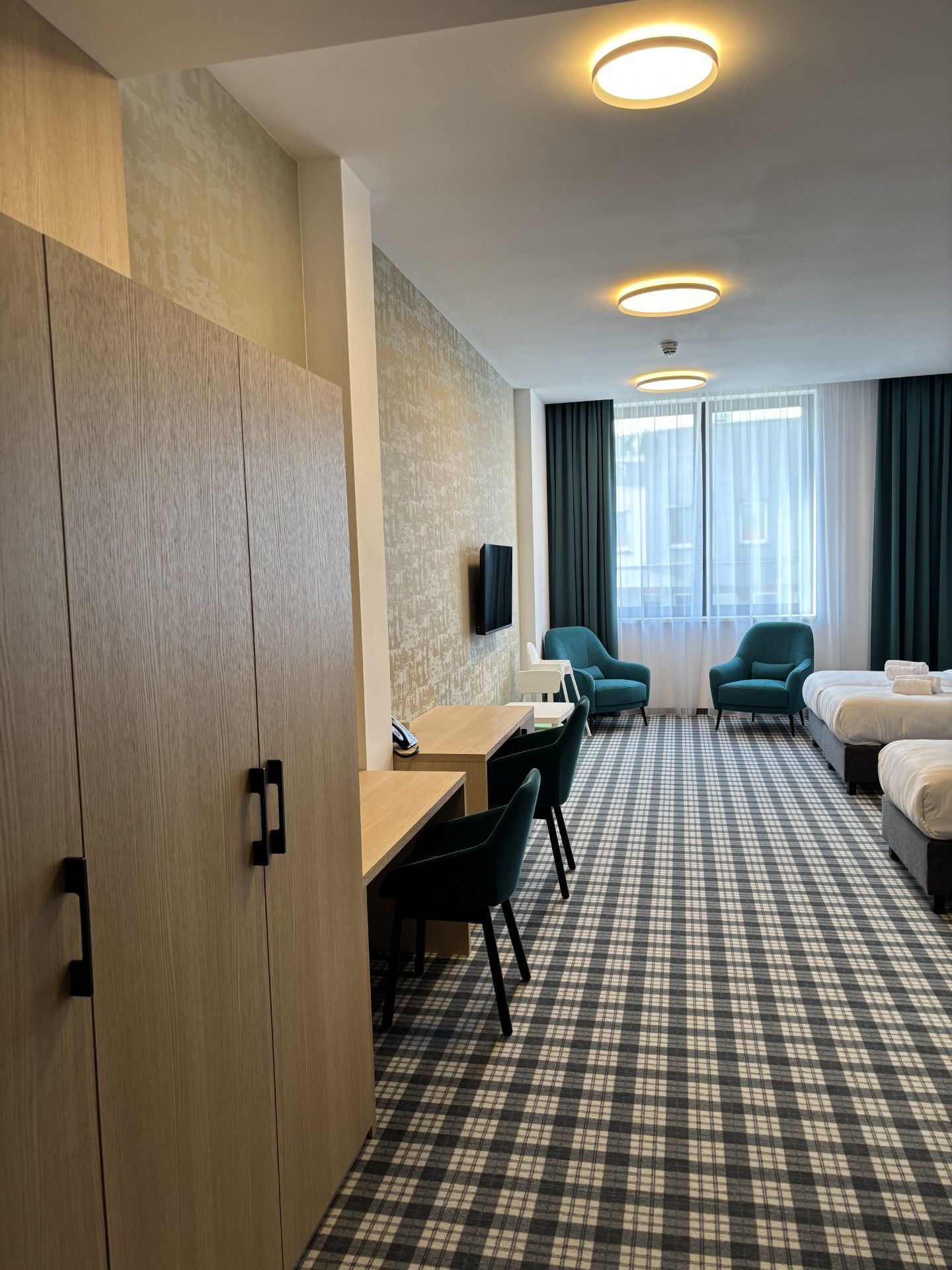 A hotel room with a checkered floor, a desk with chairs, a bed, and a TV mounted on the wall.