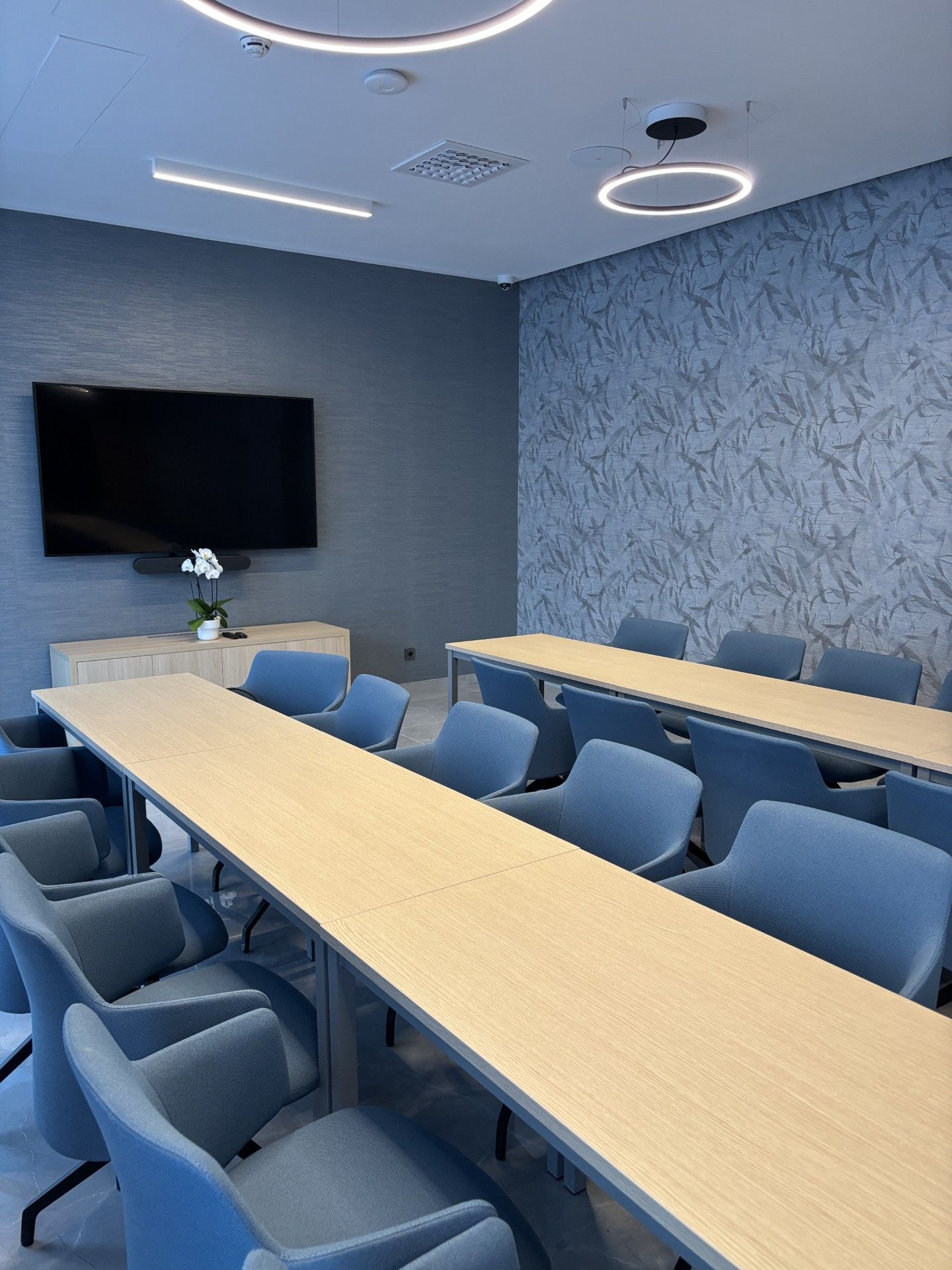 Un salón de conferencias con una larga mesa, sillas azules, un televisor de pantalla plana y un papel tapiz con diseño.