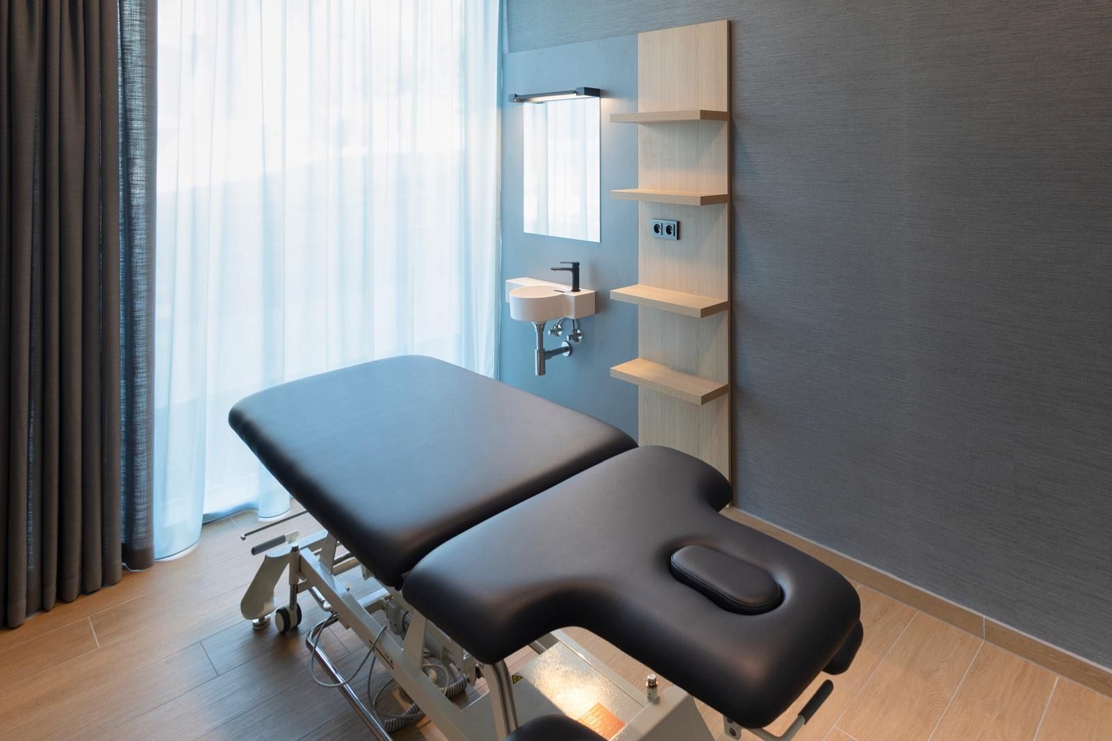 Una camilla de exploración médica con reposapiés, un estante de pared y un ventanal con cortinas.
