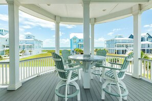 Et overdækket udendørsområde med et rundt bord og stole, med udsigt til en strandpromenade med flere huse.