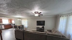 Uma sala de estar com sofá seccional, ventilador de teto, televisão de tela plana e um espaço de jantar com mesa e cadeiras.