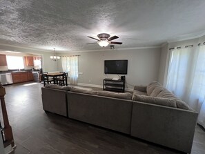 Een woonkamer met een hoekbank, een plafondventilator, een flatscreen-tv en een eethoek met een tafel en stoelen.