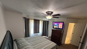 Quarto com cama, cômoda, ventilador de teto, televisão e porta.