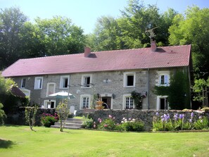 Un bâtiment en pierre avec un toit recouvert de tuiles rouges, un mur en pierre et un jardin agrémenté de fleurs et d’un petit arbre.
