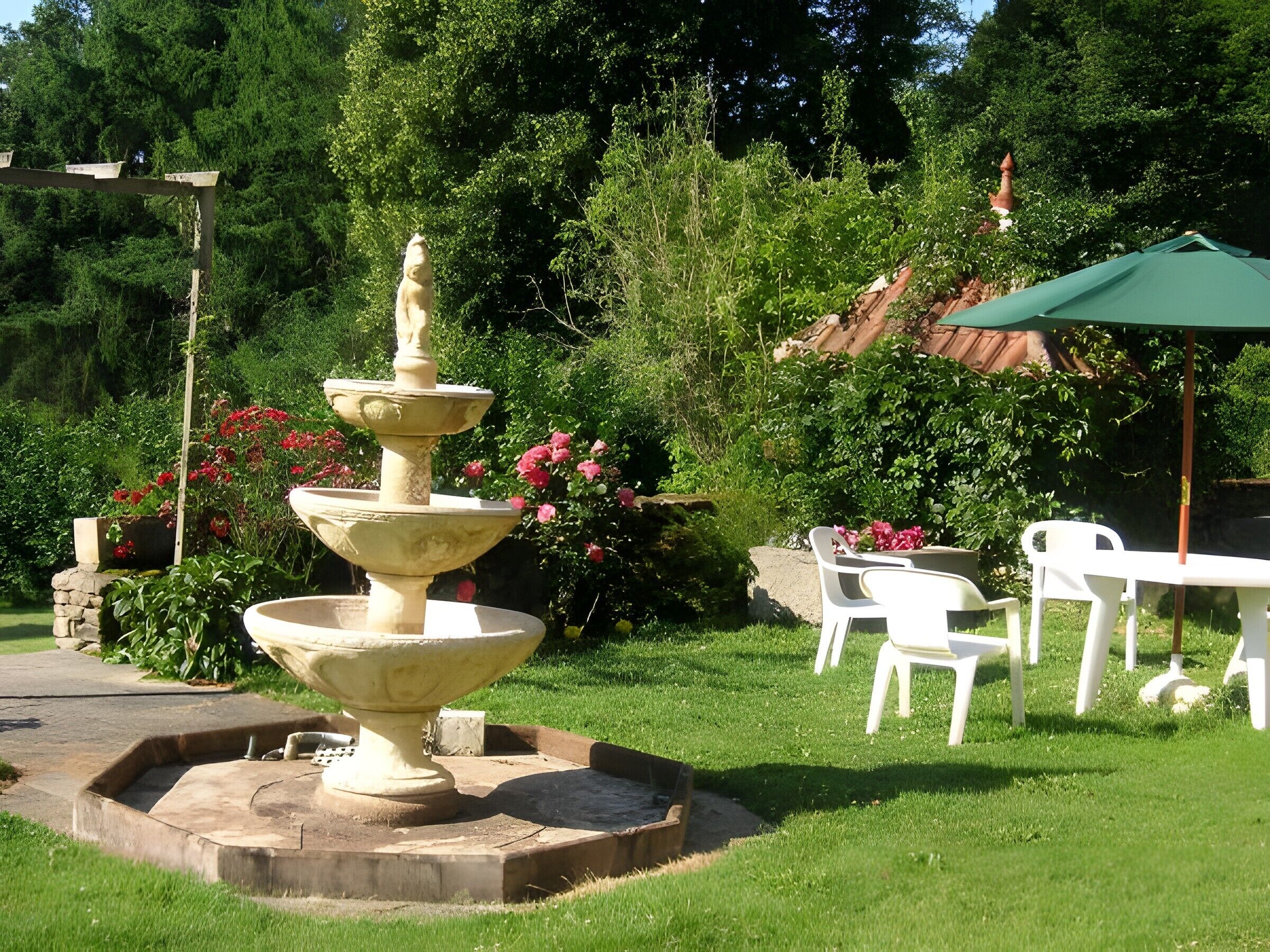 Un jardin agrémenté d’une fontaine à plusieurs niveaux, de chaises en plastique blanches et d’un parasol vert.