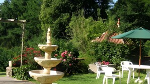 A garden with a multi-tiered fountain, white plastic chairs, and a green umbrella.
