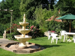 Un jardin agrémenté d’une fontaine à plusieurs niveaux, de chaises en plastique blanches et d’un parasol vert.