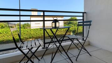 Een balkon met een metalen tafel en stoelen, een kleine lantaarn, en uitzicht op groen en gebouwen.