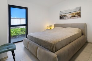 A bedroom with a bed, a chair, a nightstand with a lamp, a window with a view of greenery, and a painting on the wall.
