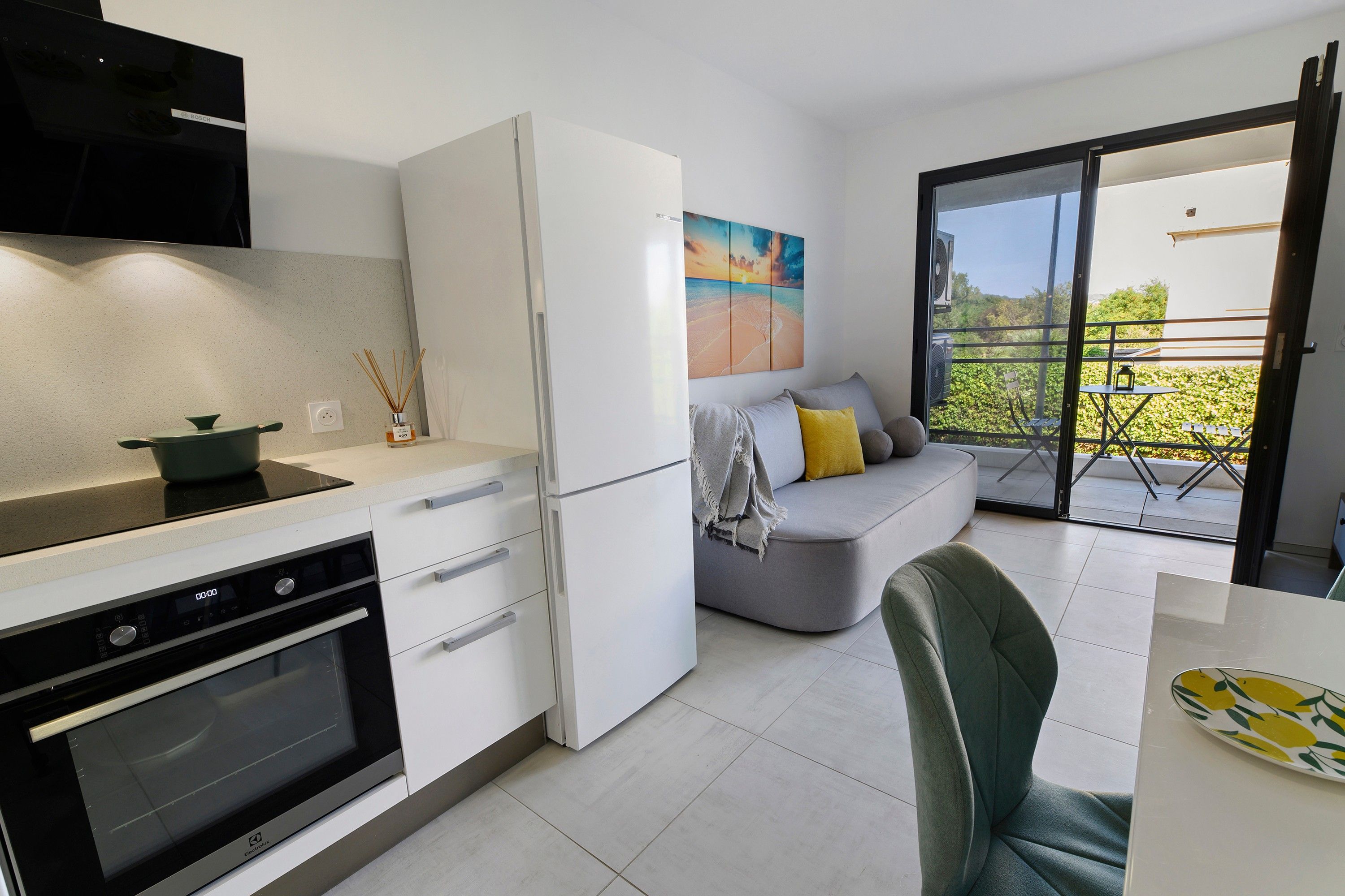 Een moderne keuken met een inbouwoven en een koelkast. Er is een zithoek met een tafel en een stoel. Een balkon met uitzicht op groen is zichtbaar.