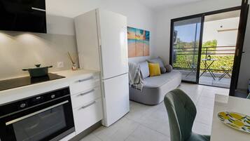 Een moderne keuken met een inbouwoven en een koelkast. Er is een zithoek met een tafel en een stoel. Een balkon met uitzicht op groen is zichtbaar.