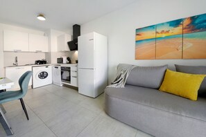Eine moderne Küche mit weißen Geräten, ein Essbereich mit Tisch und Stühlen sowie ein Wohnbereich mit Sofa und einem Strandbild.