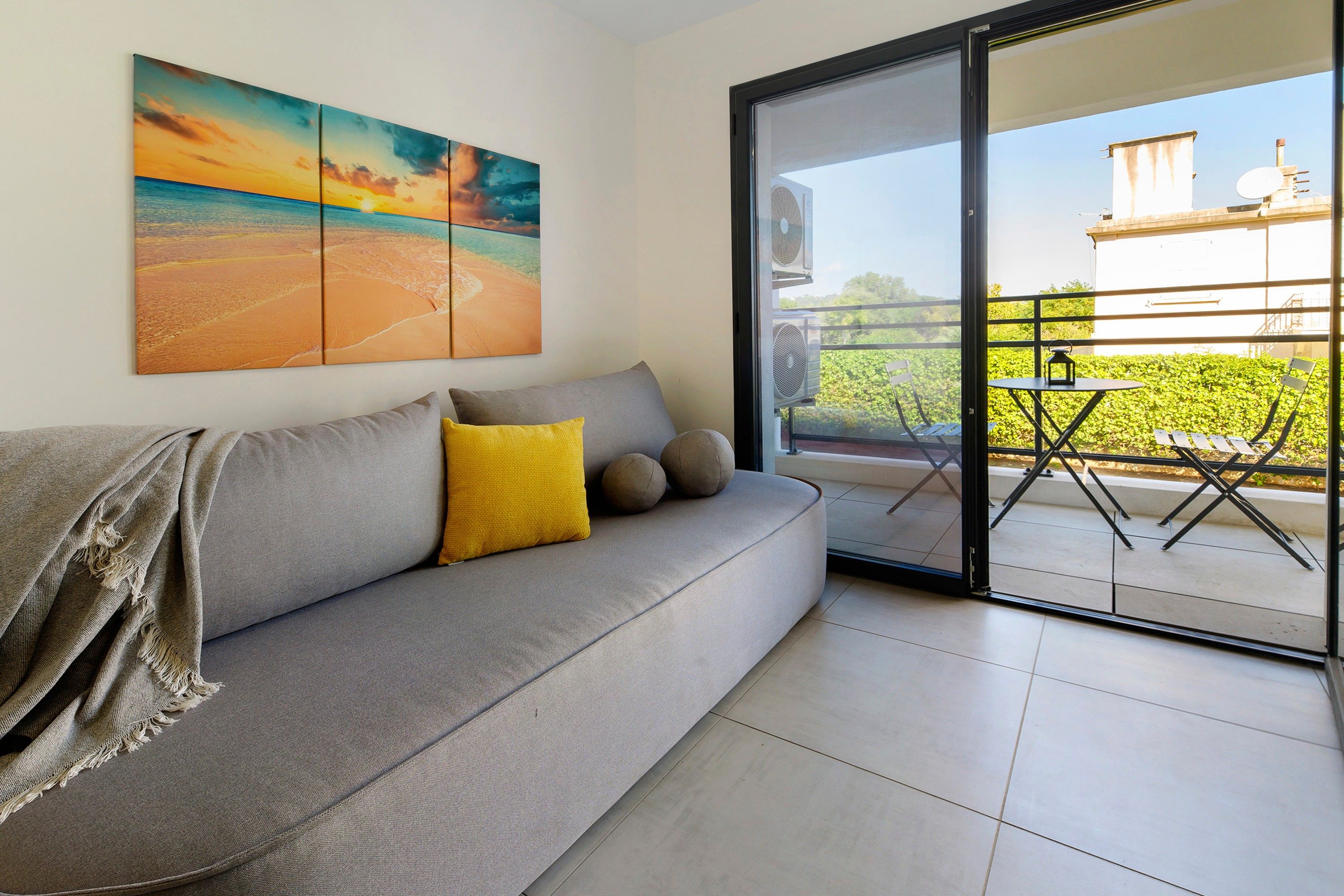 Een moderne woonkamer met een grijze bank, een geel kussen en een groot schilderij van een strand.