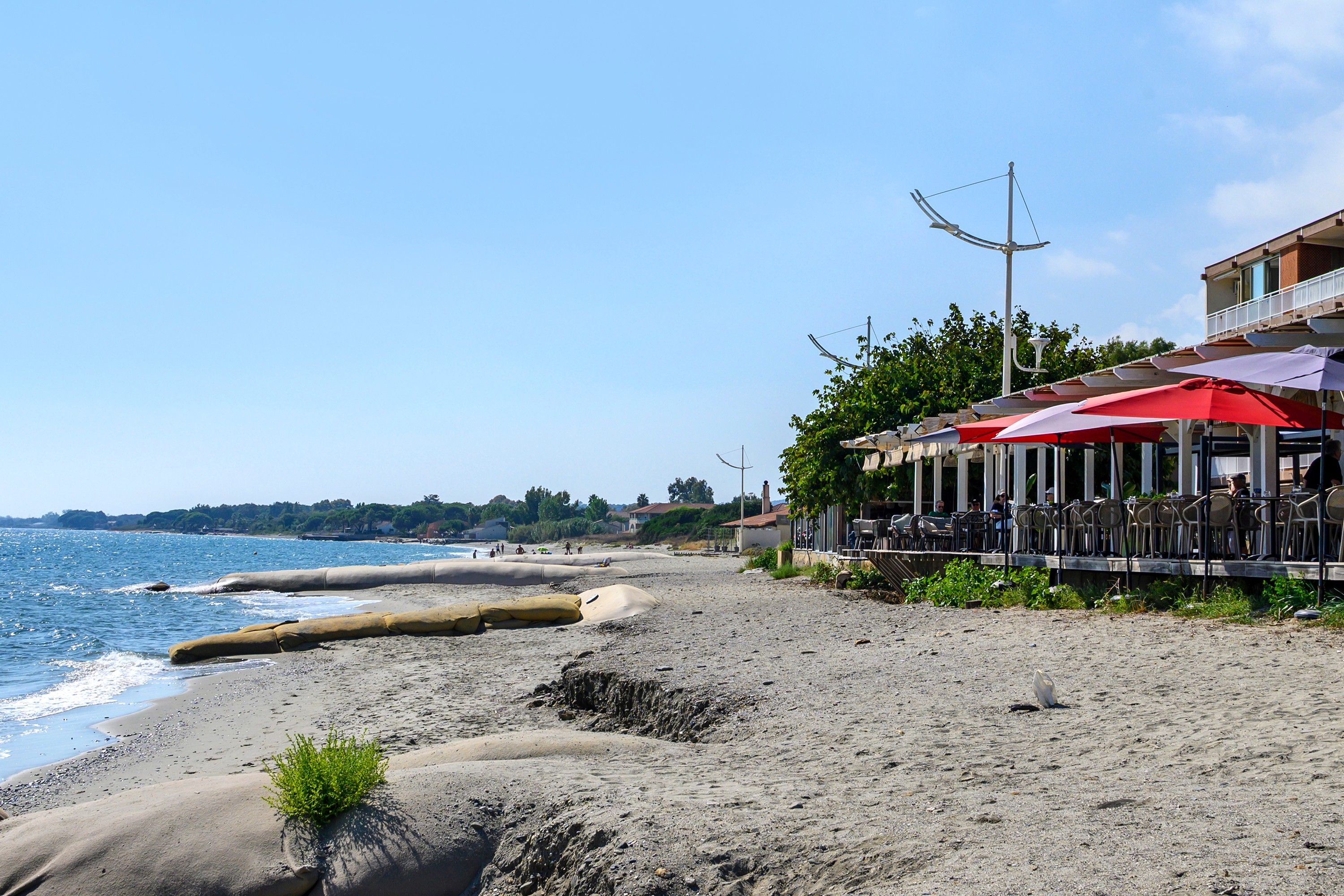 Eine Küstenlandschaft mit Sandstrand, einem Restaurant mit Außenterrasse und freiem Blick auf das Meer.