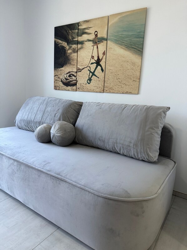 Un salon moderne avec un canapé gris, deux coussins gris et un grand tableau représentant une ancre sur une plage.