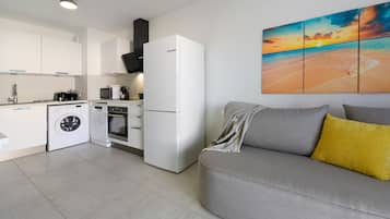 Een moderne keuken met witte apparaten, een wasmachine, en een zithoek met een grijze bank en een geel kussen.