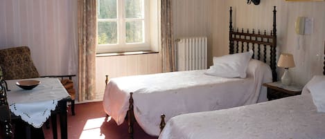 Une chambre avec deux lits, une table avec un bol, une chaise, une fenêtre avec des rideaux, un radiateur et un téléphone fixé au mur.