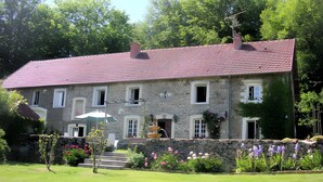 A stone building with a red-tiled roof, a stone wall, and a garden with flowers and a small tree.