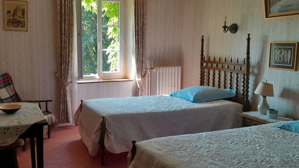 Quarto com duas camas, uma mesa com uma tigela, uma cadeira, uma janela com cortinas e um radiador.