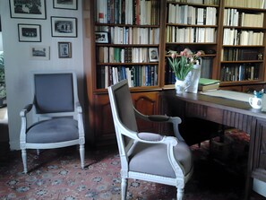A room with two white chairs, a wooden desk, a bookshelf filled with books, and a patterned carpet.