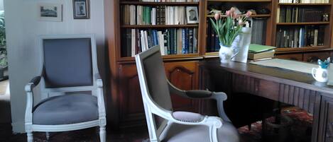 Une pièce avec deux chaises blanches, un bureau en bois, une bibliothèque remplie de livres et un tapis à motifs.