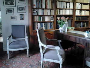 A room with two white chairs, a wooden desk, a bookshelf filled with books, and a patterned carpet.