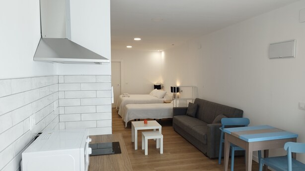 A compact hotel room with a kitchenette, two beds, a sofa, and a small table with chairs.