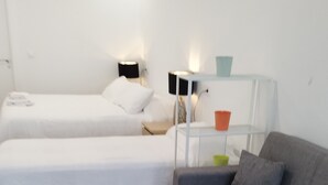 A room with two beds, a grey sofa, a small white table, and a nightstand with lamps.