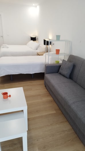 Una habitación con dos camas, un sofá gris, una mesita blanca y una mesita de noche con lámparas.