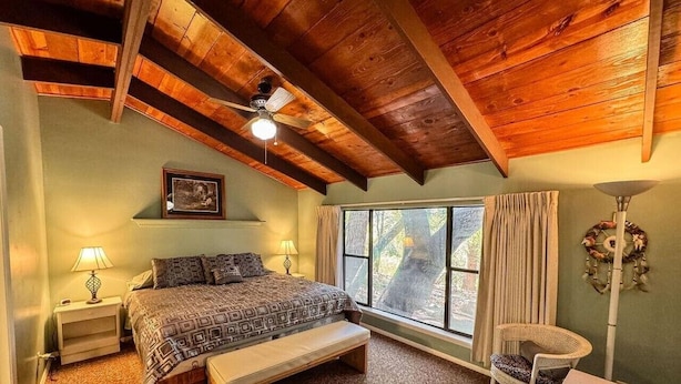 Une chambre à coucher comprenant un lit, un banc, des tables de chevet, une fenêtre avec des rideaux, un ventilateur de plafond et un luminaire fixé au mur.