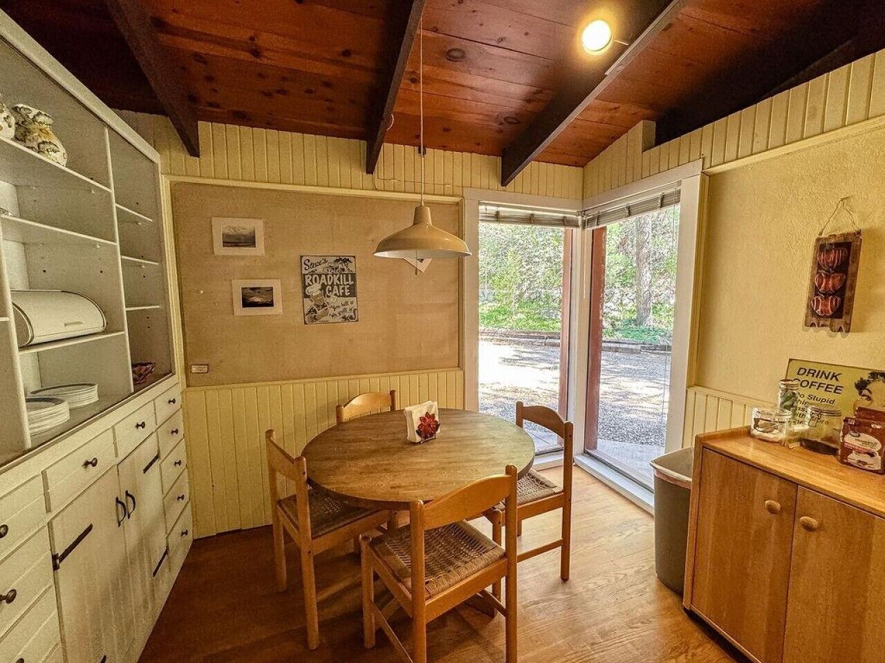 A cozy dining area with a wooden table and chairs, a hanging pendant light, and a view of the outdoors through a glass door.