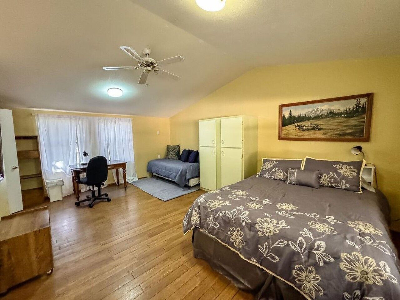 A bedroom with a bed, desk, chair, wardrobe, and a painting on the wall.