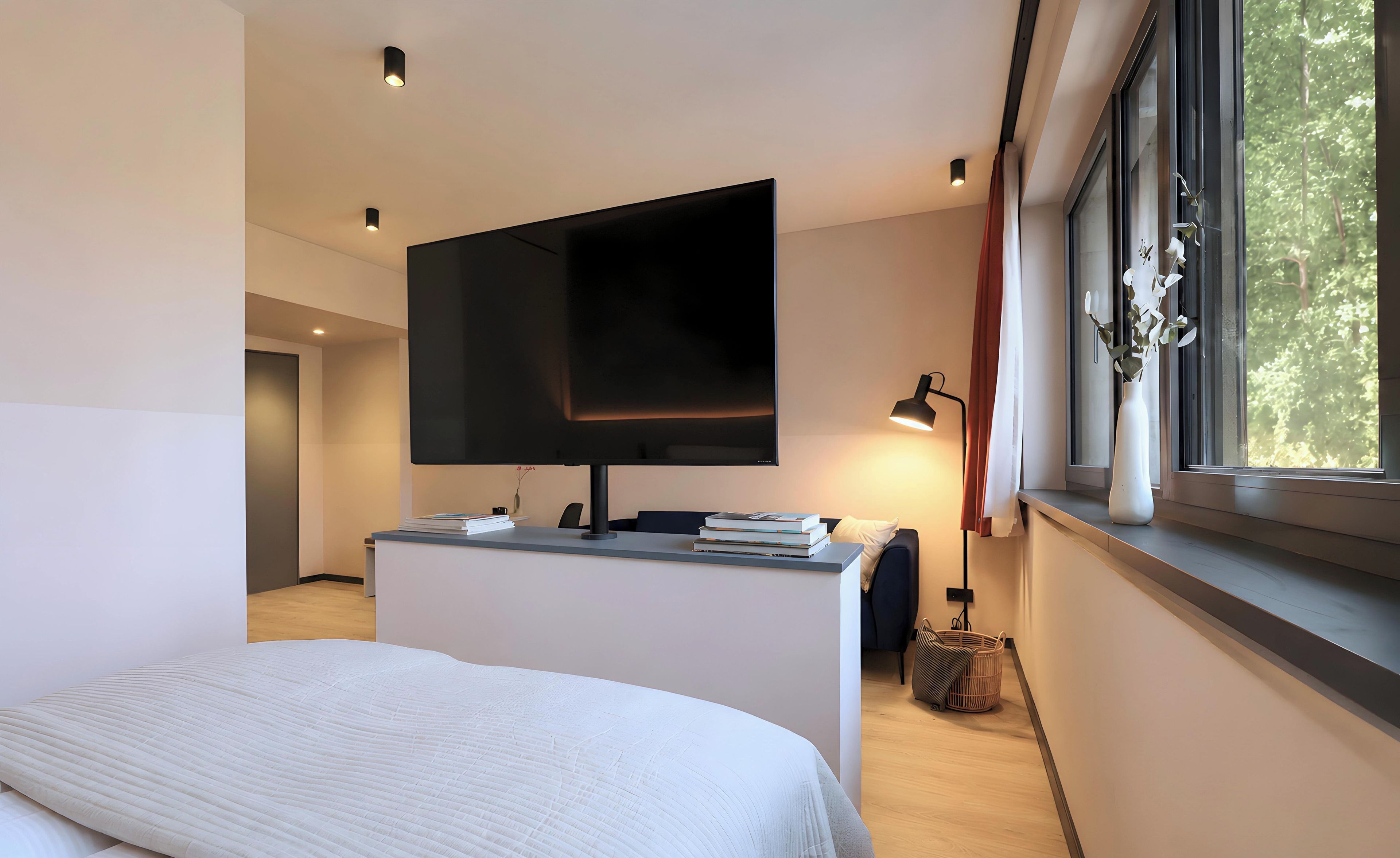 A modern bedroom with a bed, a flat-screen TV mounted on a wall, a desk with books, a lamp, and a window with a view of trees.