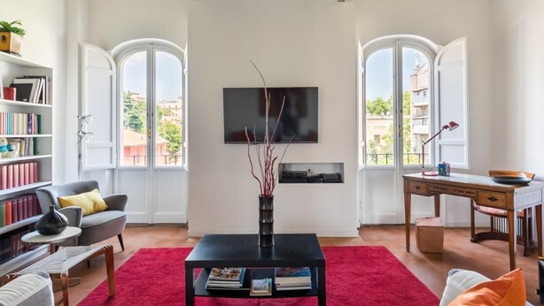 A living room with a red carpet, a black coffee table, a bookshelf, a grey sofa, a TV mounted on the wall, and two windows with arched tops.