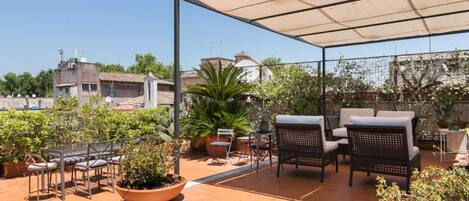 A covered outdoor seating area with chairs and tables, surrounded by potted plants and a clear sky.