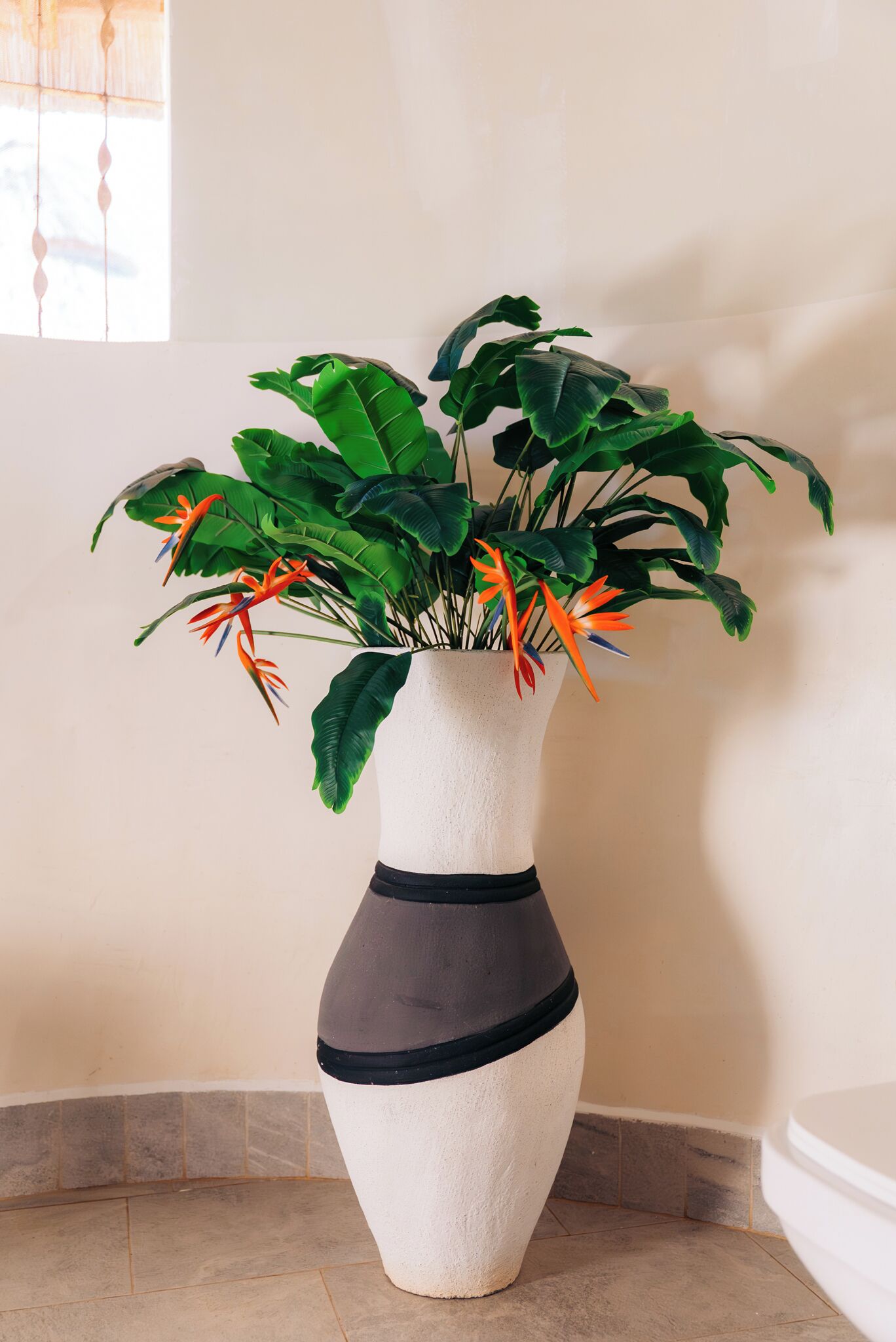 Ein Blumentopf mit einer breiten, grünen Belaubung und orangefarbenen Blüten in einer weißen Vase mit schwarzen Bändern.