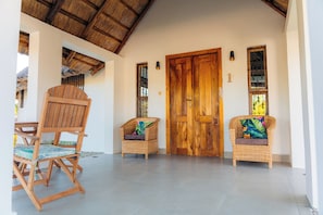 A room with a wooden door, two wicker chairs with colorful cushions, a wooden chair, and a thatched roof visible through an open doorway.
