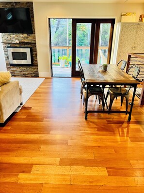 Ein geräumiger Wohnbereich mit Holzboden, einer Couch, einem Esstisch mit Stühlen, einem an einer Steinwand montierten Flachbildfernseher und einer Glastür, die zu einer Außenpatio führt.