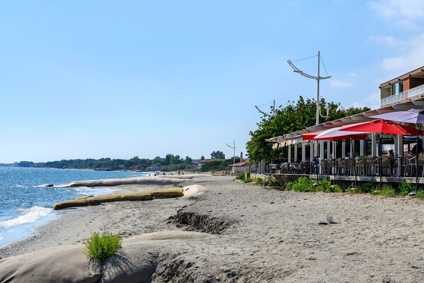 A coastal area with a sandy beach, a restaurant with outdoor seating, and a clear view of the sea.