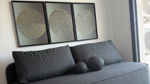 A modern grey sofa with three cushions in a minimalist living room with a large window and three framed abstract artworks on the wall.