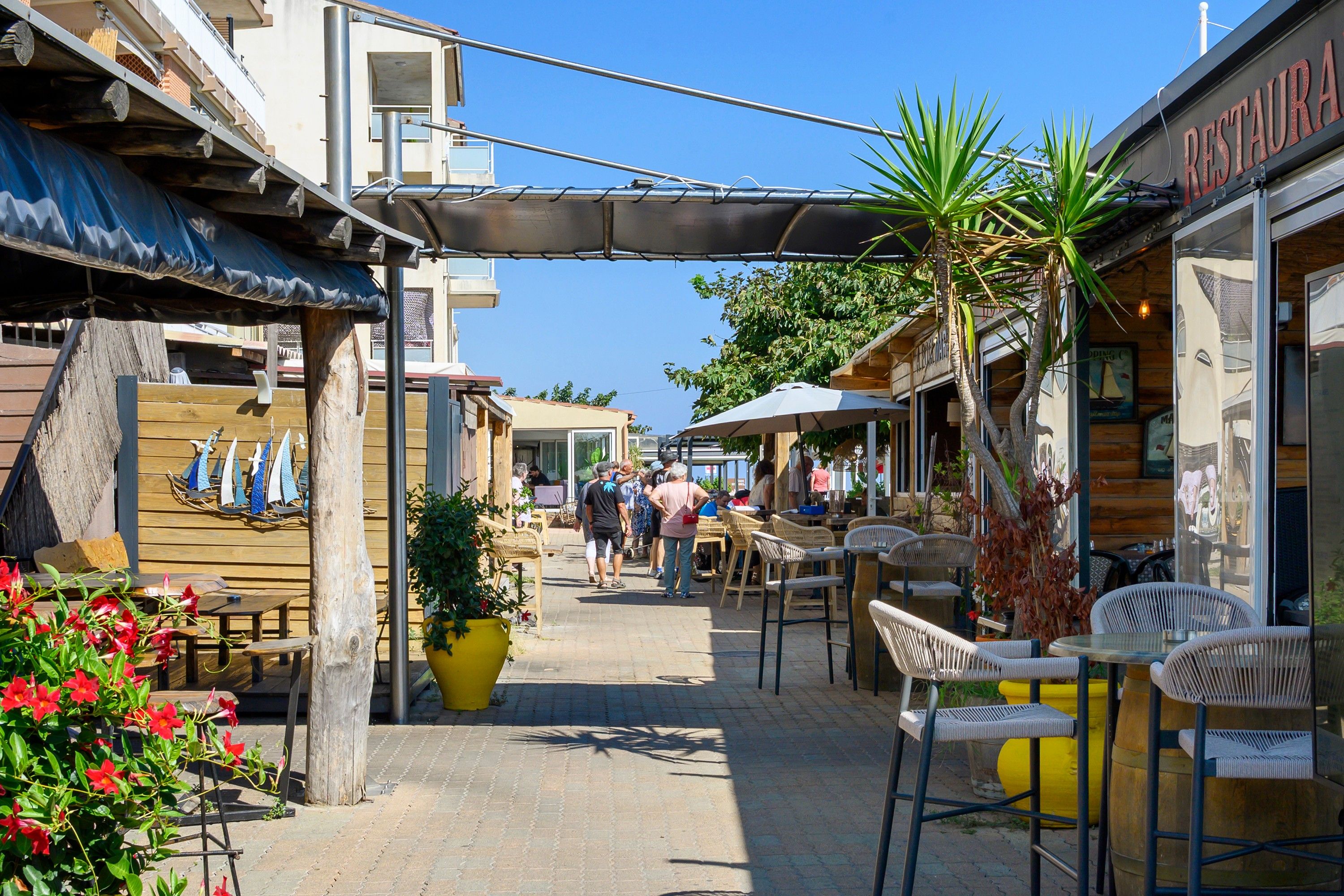 A narrow outdoor corridor with outdoor seating, potted plants, and a variety of shops and eateries.