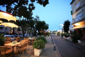 Un espace extérieur aménagé avec des tables, des chaises, des parasols et des plantes en pot, le long d’une rue.