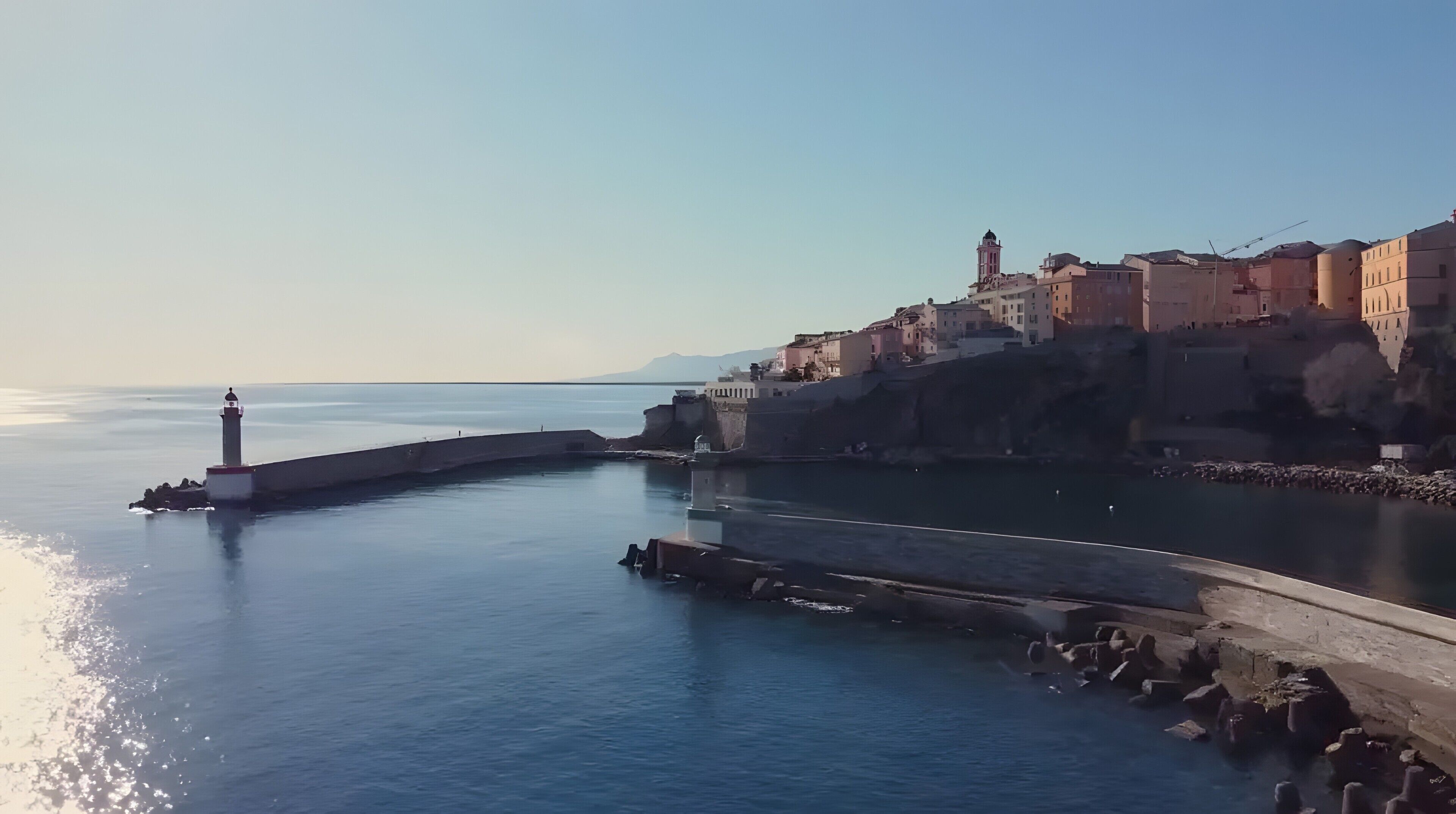 Un pueblo costero con un faro destacado, un mar tranquilo y edificios a lo largo del paseo marítimo.